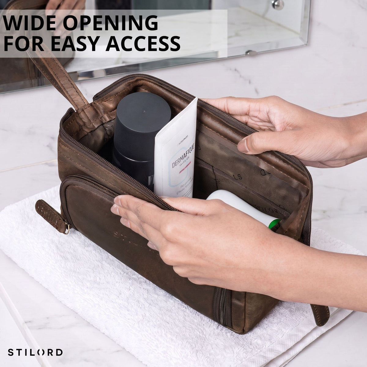 Brown leather Henry toiletry bag with wide opening, hands showing interior on marble sink, STILORD embossing visible